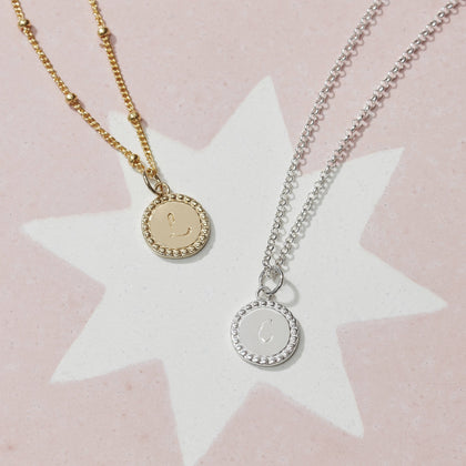 a gold friendship disc charm on a gold bead chain necklace lying next to a silver friendship disc charm on a silver belcher chain necklace